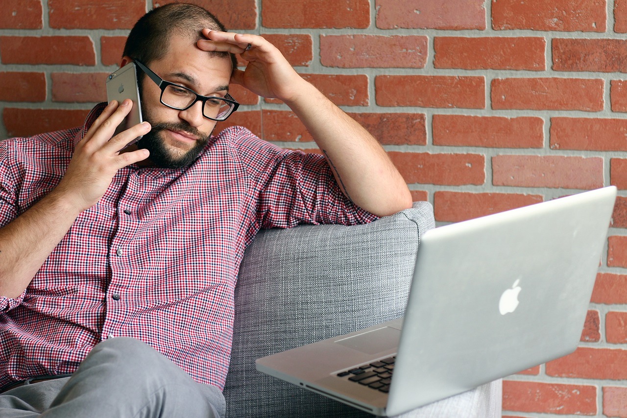 A man on his phone and in front of his laptop looking frustrated.